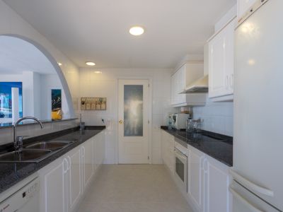 Cocina funcional con mobiliario blanco, encimeras de granito oscuro y electrodomésticos integrados, conectada a la zona de estar mediante un arco.