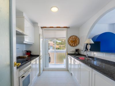 Cocina tipo galería con muebles blancos, encimeras de granito oscuro y azulejos. Incluye horno, nevera y lavadora en el lavadero adyacente, con vistas a la ladera.