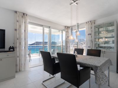 Comedor luminoso con mesa moderna y sillas, acceso directo a terraza acristalada con vistas panorámicas al mar y al Peñón de Ifach. Vitrina con cristalería.