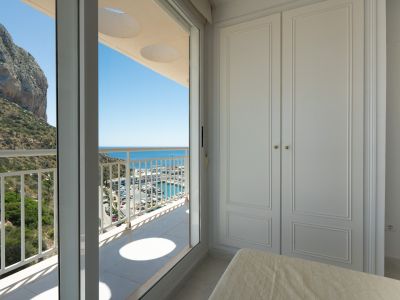 Dormitorio con armario empotrado blanco y acceso a terraza. Vistas panorámicas al Peñón de Ifach, el puerto deportivo y el mar Mediterráneo bajo un cielo azul.