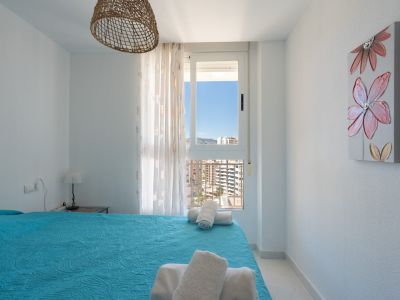 Dormitorio luminoso con cama de edredón azul y toallas blancas. Dispone de una ventana con vistas urbanas y montañosas, lámpara de techo de mimbre y un cuadro floral.