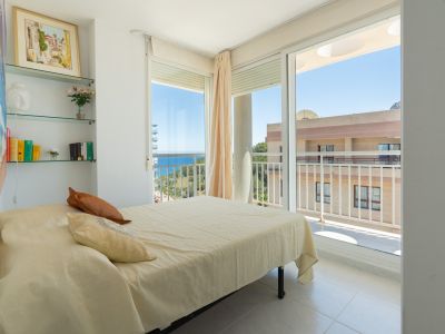 Dormitorio luminoso con cama de matrimonio, estanterías de cristal decoradas y acceso directo a una terraza con vistas parciales al mar y al entorno urbano.