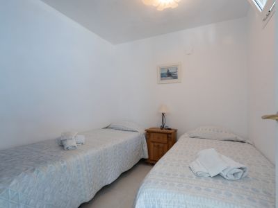 Dormitorio secundario con dos camas individuales y colchas estampadas. Incluye mesita de noche de madera, lámpara y cuadro decorativo, todo sobre suelo de azulejos claros.