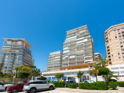 Edificios residenciales modernos con balcones de cristal y uno de fachada tradicional, junto a una calle con palmeras y vehículos aparcados bajo un cielo azul claro.