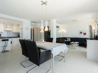 Espacio diáfano con cocina blanca, barra americana, comedor con lámpara colgante de tres luces y salón con sofá seccional negro.