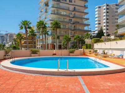La piscina comunitaria, con sus aguas azules y escaleras de acero inoxidable, está rodeada por un solárium de baldosas de terracota y palmeras, con edificios residenciales al fondo.