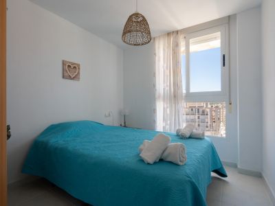 Luminoso dormitorio con cama doble vestida en turquesa, lámpara colgante de fibra natural y ventana con vistas urbanas y edificios circundantes.