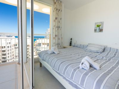 Luminoso dormitorio con cama doble y ropa de cama a rayas, mesita de noche con lámpara y acceso directo a balcón privado con vistas al mar y urbanas.