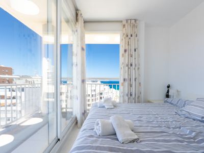 Luminoso dormitorio con cama doble y ropa de cama a rayas, ofreciendo acceso directo a una terraza con vistas panorámicas al mar y al cielo azul.