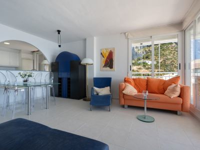 Luminoso salón-comedor con suelo de baldosas claras, mesa de cristal y sofá naranja. Amplias puertas correderas con vistas a la ladera verde.