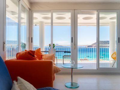 Luminoso salón con sofá naranja y mesa de cristal, con amplias puertas correderas que revelan una terraza y vistas directas al mar, barcos y montañas lejanas.