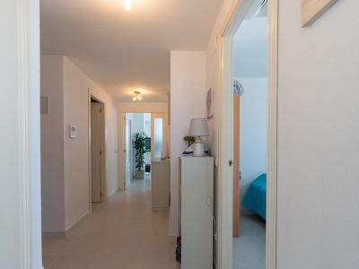 Pasillo interior con suelo de baldosas claras y paredes blancas. Varias puertas conducen a estancias, incluyendo un dormitorio visible a la derecha y una sala de estar al fondo.