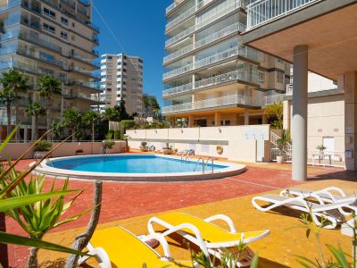 Piscina comunitaria de forma circular rodeada por una terraza pavimentada en tonos rojizos y amarillos, con tumbonas y palmeras bajo modernos edificios residenciales.
