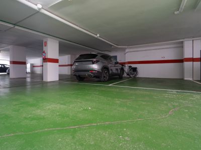 Plaza de aparcamiento subterráneo con suelo verde, pilares blancos con franjas rojas, un SUV gris y una bicicleta. Extintor visible.