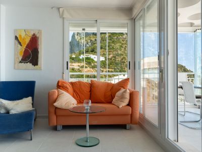 Salón luminoso con sofá naranja y sillón azul, ofreciendo vistas a la montaña a través de amplias puertas correderas que dan a una terraza con mobiliario exterior y techo decorativo.