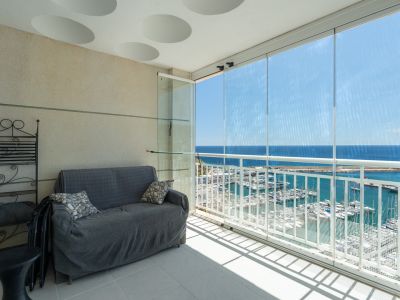 Terraza acristalada con vistas frontales al puerto deportivo y al mar, equipada con sofá y estantería de hierro forjado, bajo un techo con detalles circulares.