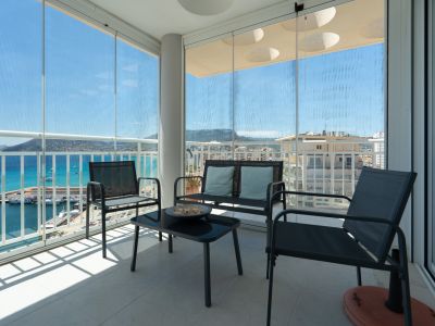 Terraza cubierta con cortinas de cristal correderas, mobiliario de exterior y vistas panorámicas al puerto deportivo de Calpe, el mar y las montañas. El suelo es de baldosas claras.