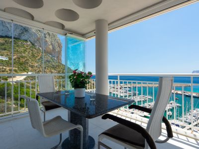 Terraza cubierta con mesa de granito y sillas modernas, equipada con cortinas de cristal, que ofrece vistas panorámicas al Peñón de Ifach y al puerto de Calpe.