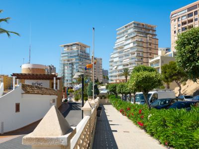 Vista del área del puerto de Calpe, con modernos edificios residenciales, un paseo adoquinado con arbustos verdes y flores rojas, y el edificio RCNC bajo un cielo azul claro.