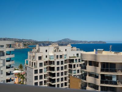 Vista panorámica al mar desde una terraza de altura, destacando la arquitectura costera moderna, aguas turquesas y montañas lejanas bajo un cielo azul claro.