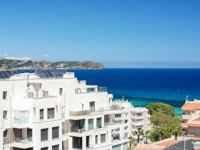 Vista panorámica de edificios residenciales blancos con balcones de cristal, paneles solares en azoteas y el azul profundo del Mediterráneo hasta la costa lejana.
