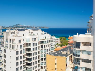 Vistas panorámicas al mar Mediterráneo y a la costa desde una planta elevada, con edificios residenciales en primer plano y montañas distantes bajo un cielo azul claro.