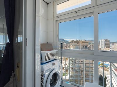Zona de lavandería funcional con lavadora y estantería, en un espacio con paredes alicatadas y amplios ventanales que enmarcan vistas urbanas a edificios, montañas y una piscina.