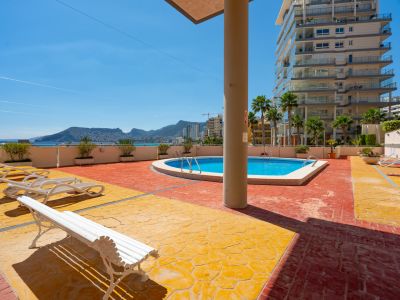 Zona de piscina comunitaria con pavimento bicolor y tumbonas. Vistas al mar Mediterráneo y las montañas, con un moderno edificio residencial al fondo.