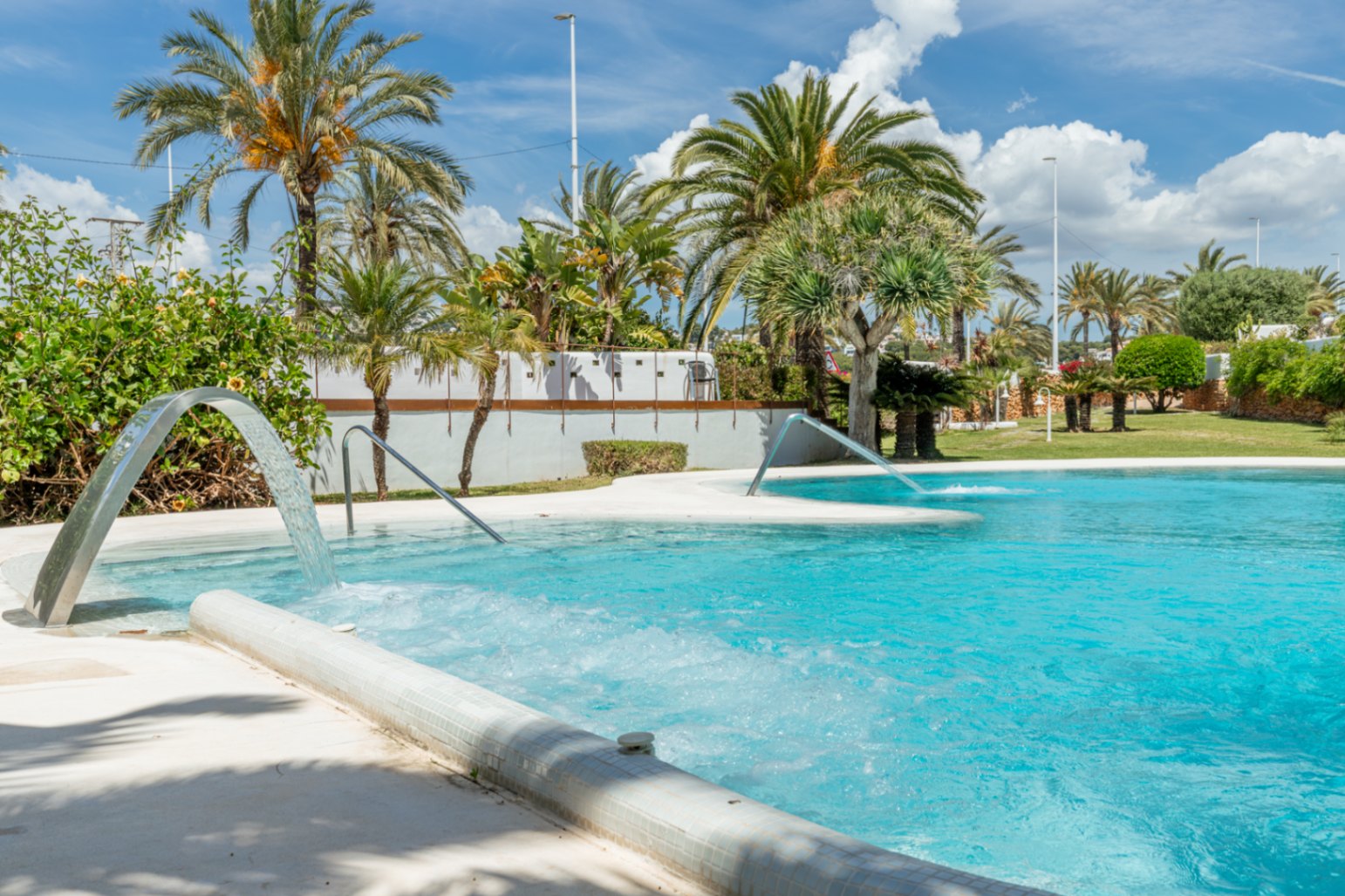 Amplia piscina comunitaria con cascadas de agua y zonas de burbujas, rodeada de exuberante vegetación y palmeras bajo un cielo azul.