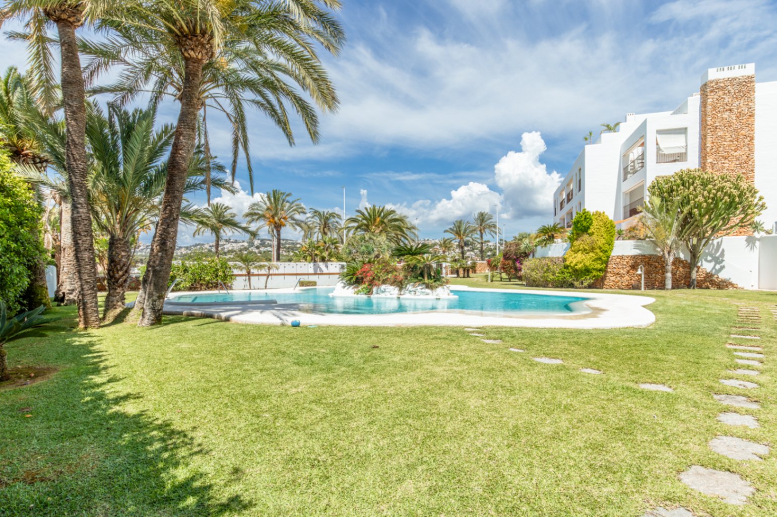 Amplia piscina comunitaria con isla ajardinada integrada, rodeada de césped impecable y palmeras maduras. El edificio residencial blanco con detalles de piedra se vislumbra bajo un cielo azul.