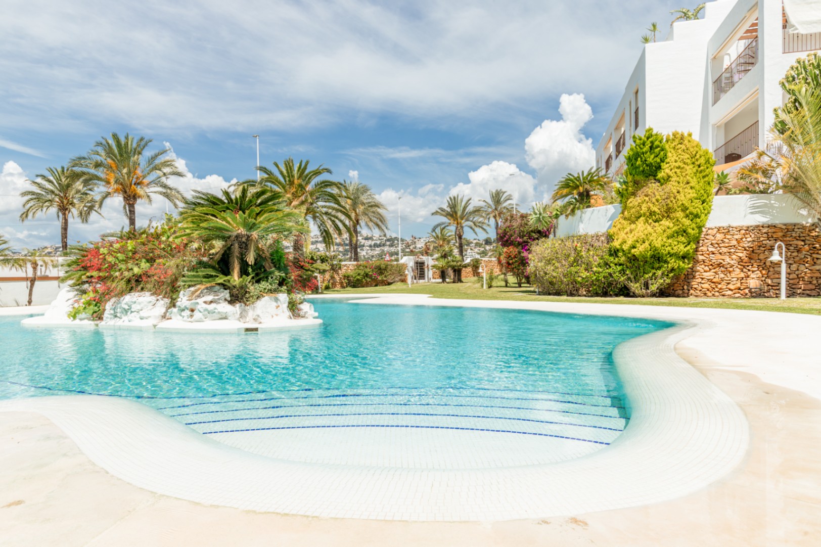 Amplia piscina comunitaria de diseño curvo con escalones de mosaico blanco y azul, realzada por una isla central con vegetación exuberante y palmeras.