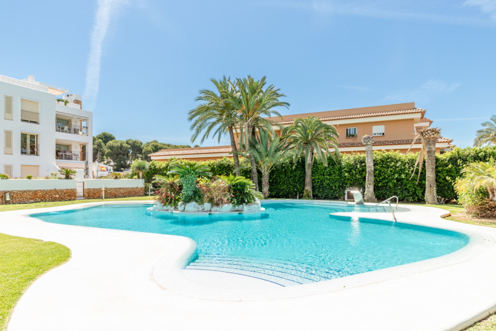Amplia piscina comunitaria de forma irregular con isla central de palmeras y vegetación exuberante, rodeada de césped y edificios residenciales bajo un cielo despejado.