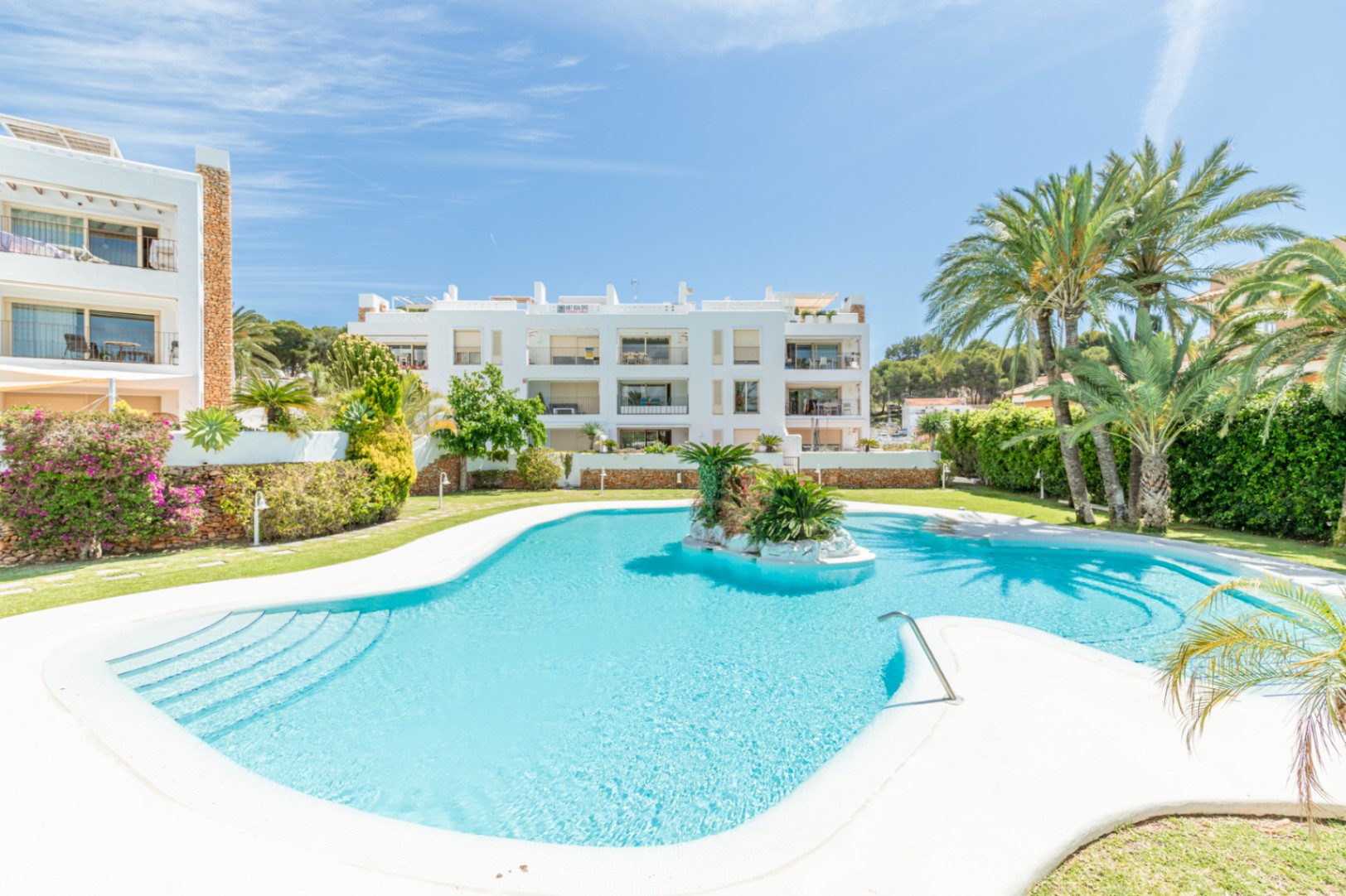 Amplia piscina comunitaria de forma libre con isla rocosa central y escaleras de acceso, rodeada de césped, palmeras y edificios residenciales blancos bajo un cielo azul.