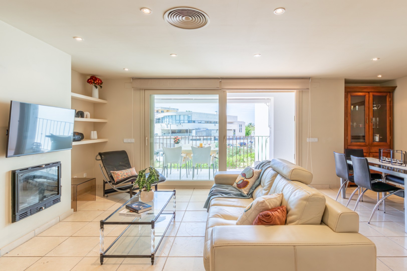 Amplia sala de estar con sofá de cuero crema, chimenea integrada y TV mural. Grandes puertas correderas conectan con una terraza privada, aportando luz natural y fluidez al espacio.