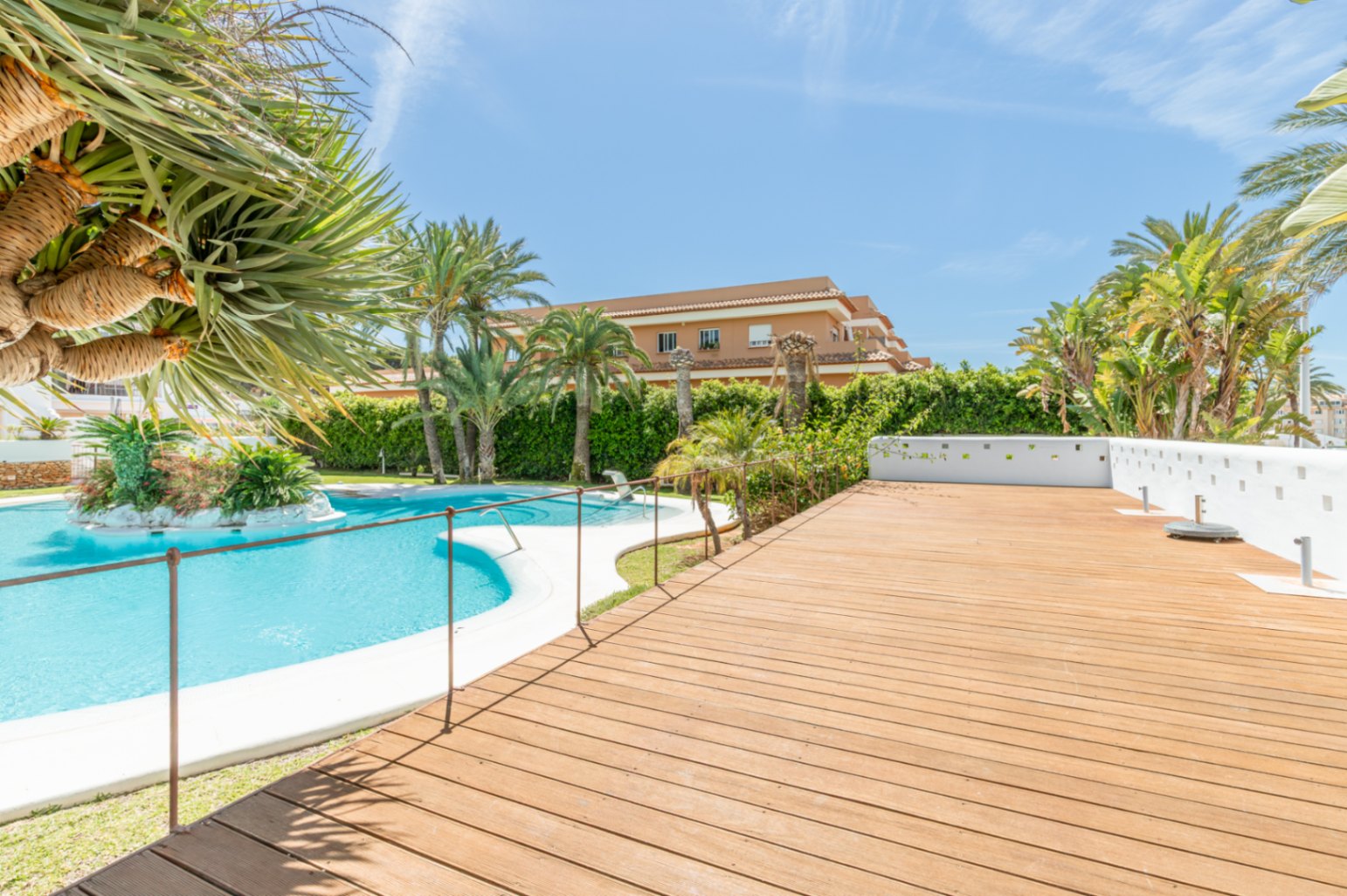 Amplia zona comunitaria con una gran piscina curva y una isla ajardinada central. La espaciosa tarima de madera adyacente está rodeada de palmeras y exuberante vegetación mediterránea.