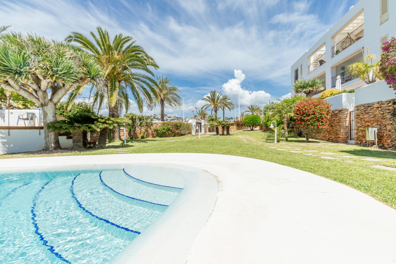 Amplia zona de piscina comunitaria con una gran alberca de escalones de mosaico azul, rodeada de césped verde y palmeras mediterráneas bajo un cielo claro.