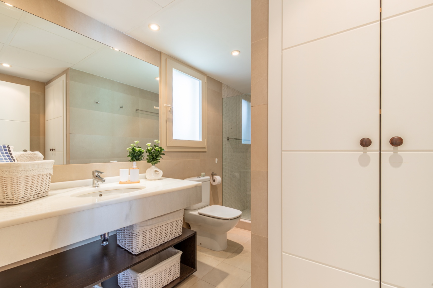 Baño completo con lavabo de mármol, amplio espejo y armarios empotrados. Incluye ducha con mampara de cristal, ventana discreta y detalles decorativos. Tonalidades neutras y gran capacidad de almacenamiento.