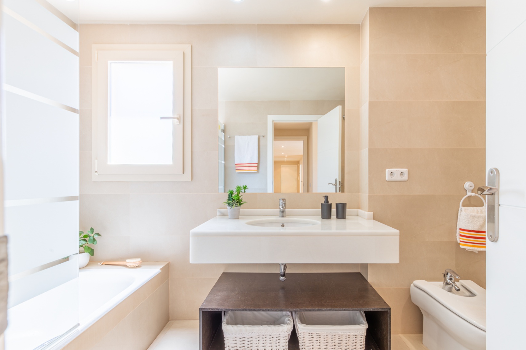 Baño completo con lavabo suspendido, encimera blanca y espejo amplio. Destacan los azulejos claros, ventana con privacidad y bañera integrada.