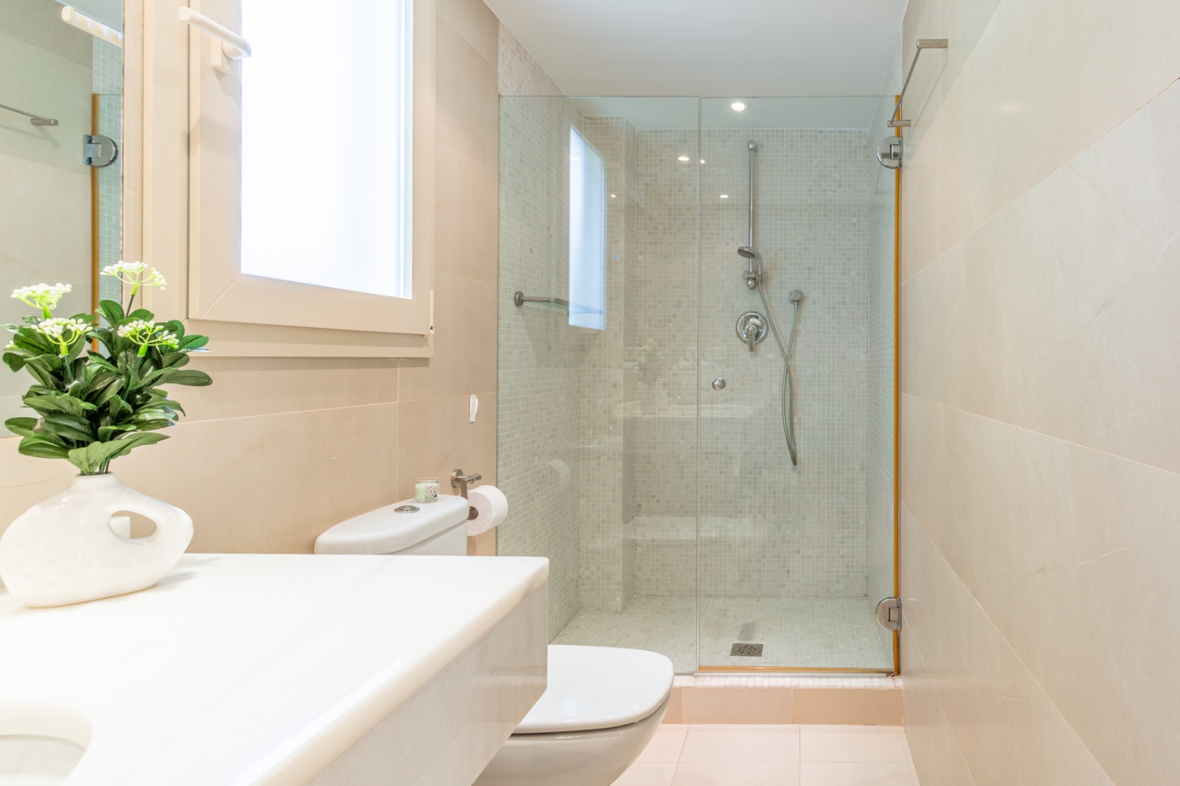 Baño moderno con inodoro, lavabo de encimera blanca y ventana. Ducha acristalada con revestimiento de mosaico y azulejos beige, complementado con luz natural.