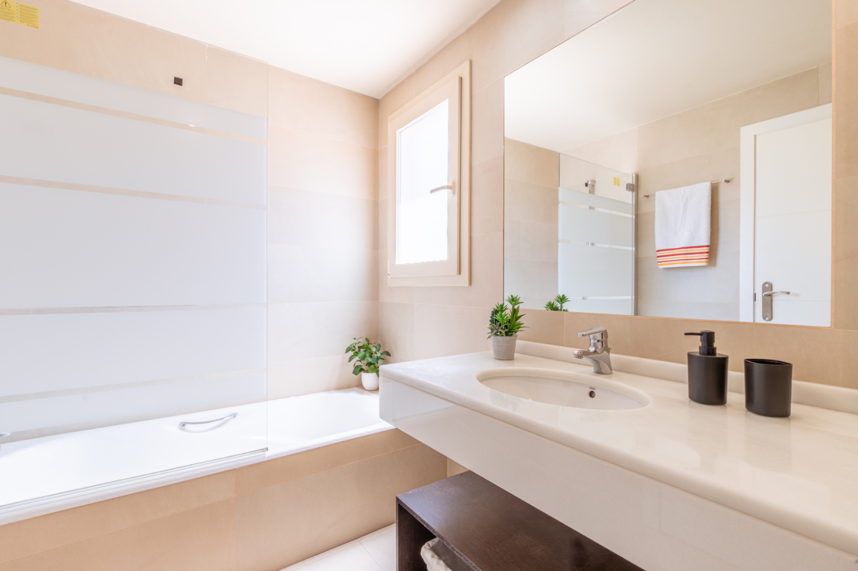 Baño moderno con lavabo flotante blanco, espejo grande y bañera con mampara de cristal a rayas, todo en un entorno de azulejos neutros.
