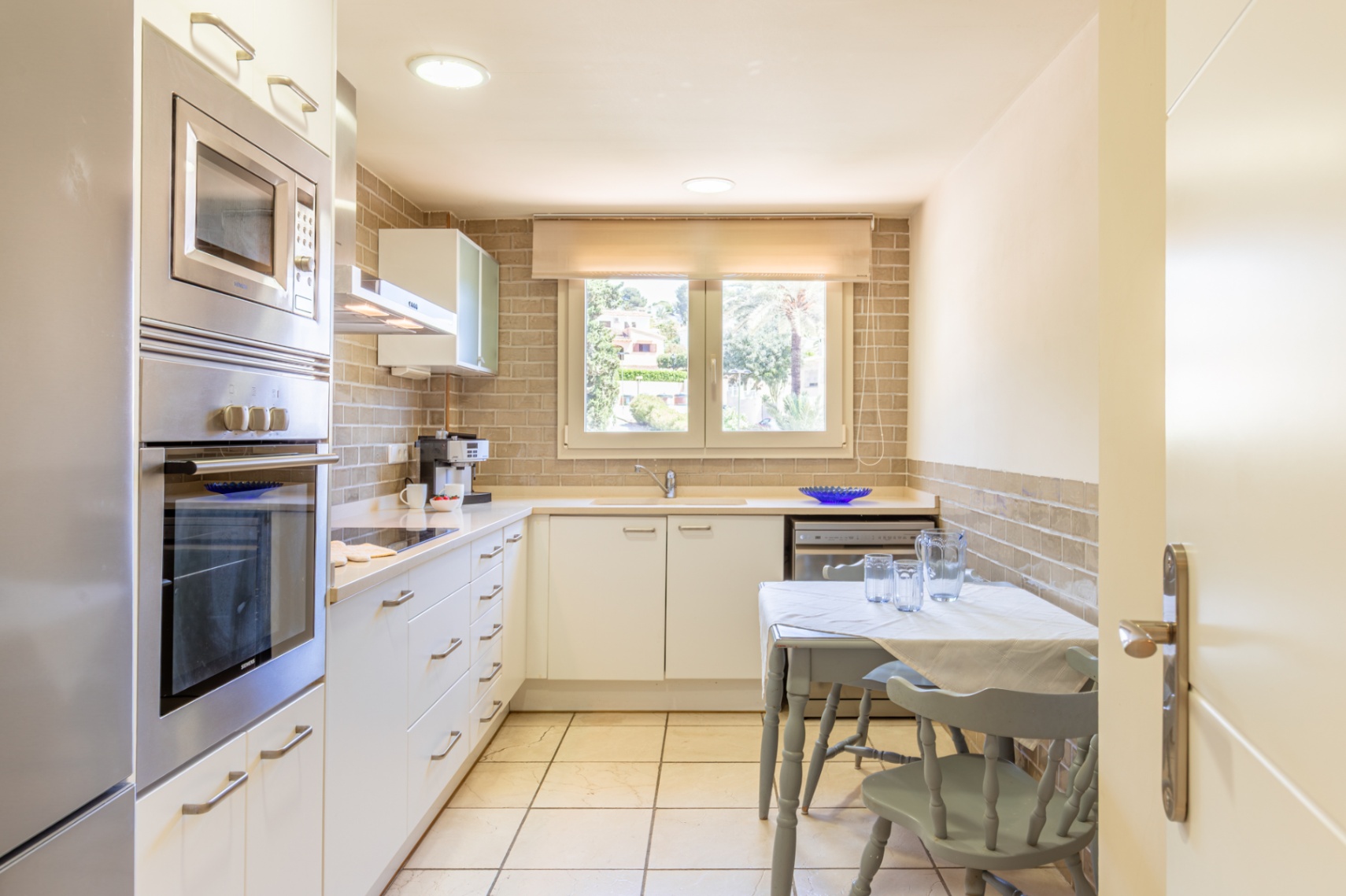 Cocina funcional con electrodomésticos de acero inoxidable integrados, encimera clara y salpicadero de ladrillo. Incluye una mesa compacta y ventana con vistas a la vegetación.