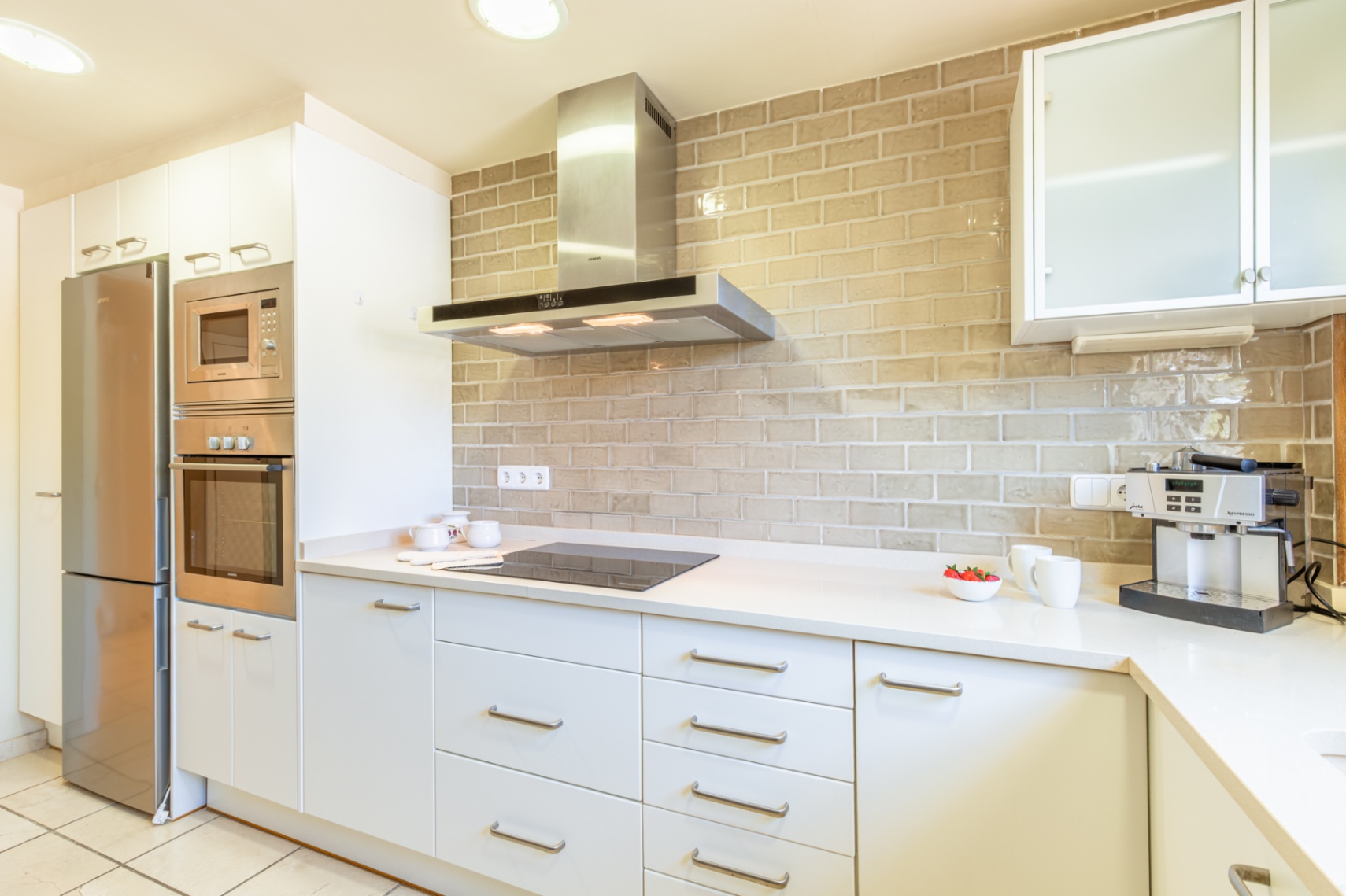 Cocina moderna con mobiliario blanco, electrodomésticos integrados de acero inoxidable y una encimera clara. Destaca el salpicadero de azulejos tipo ladrillo beige y la placa de inducción.