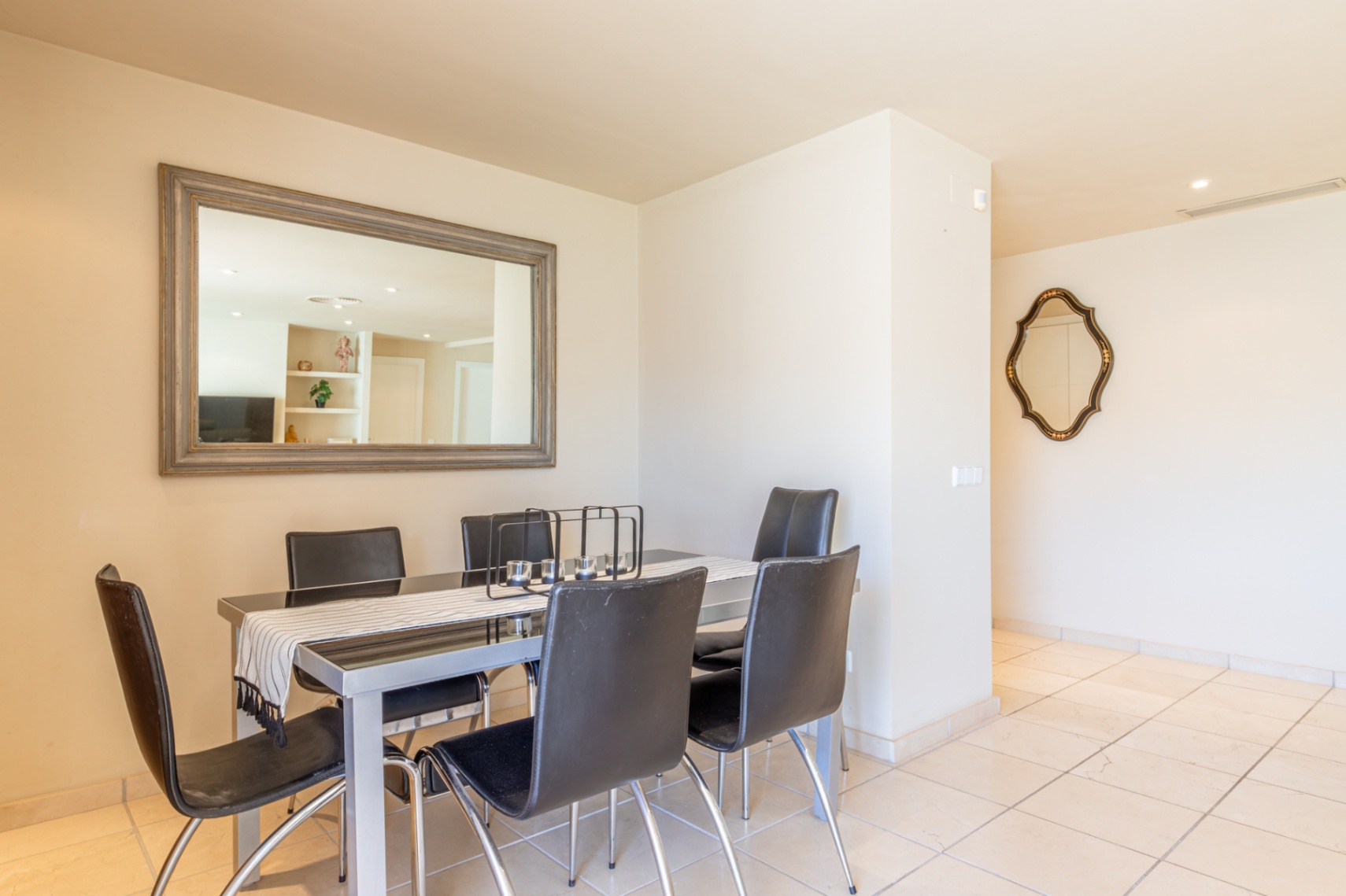 Comedor con mesa de cristal negro y seis sillas, complementado por un gran espejo con marco gris que amplía el espacio y un espejo decorativo. Suelo de baldosas claras.