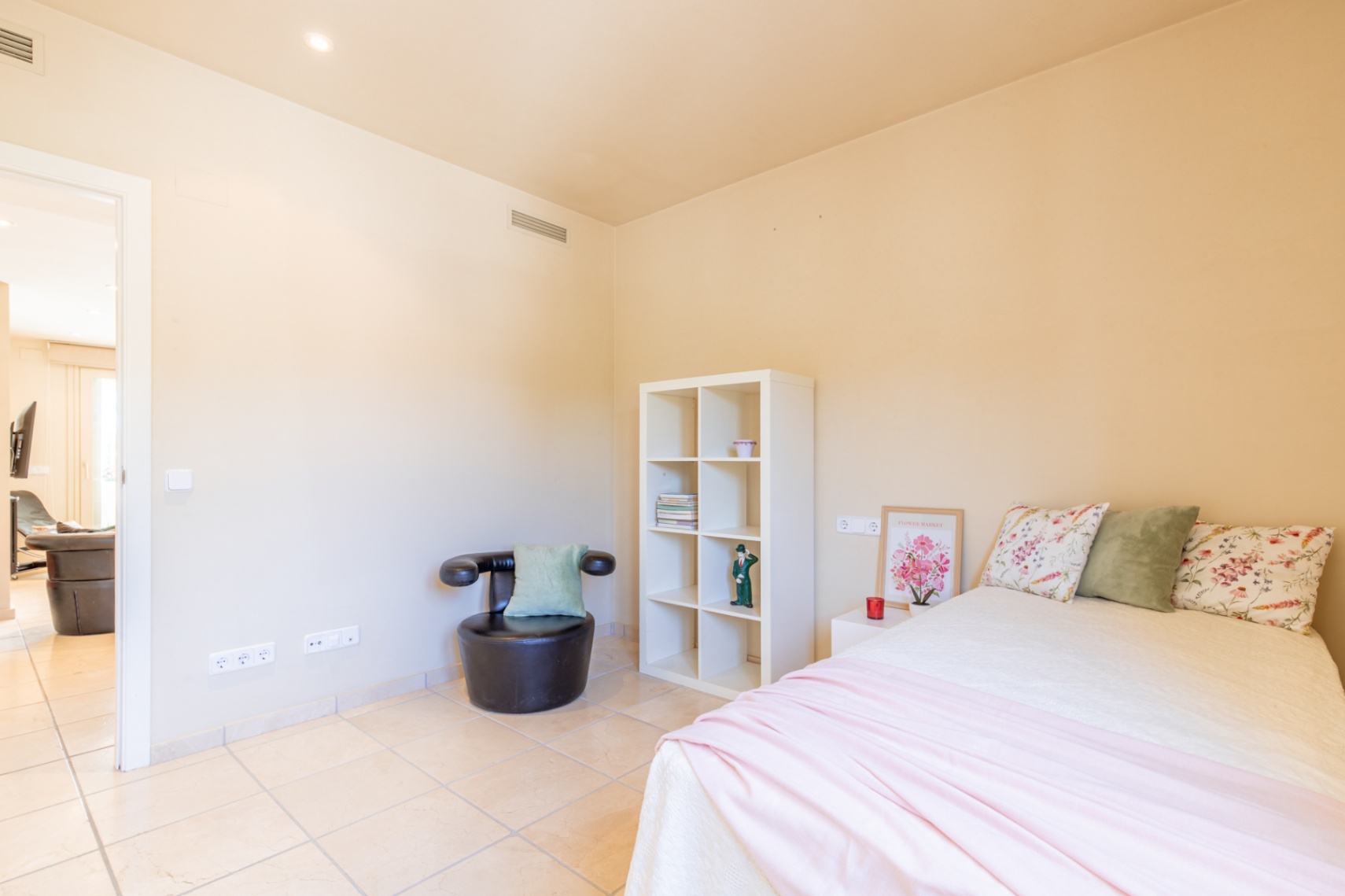 Dormitorio luminoso con cama doble, suelo de baldosas claras, paredes en tono crema y detalles decorativos. Incluye estantería blanca, sillón y acceso a otra estancia.