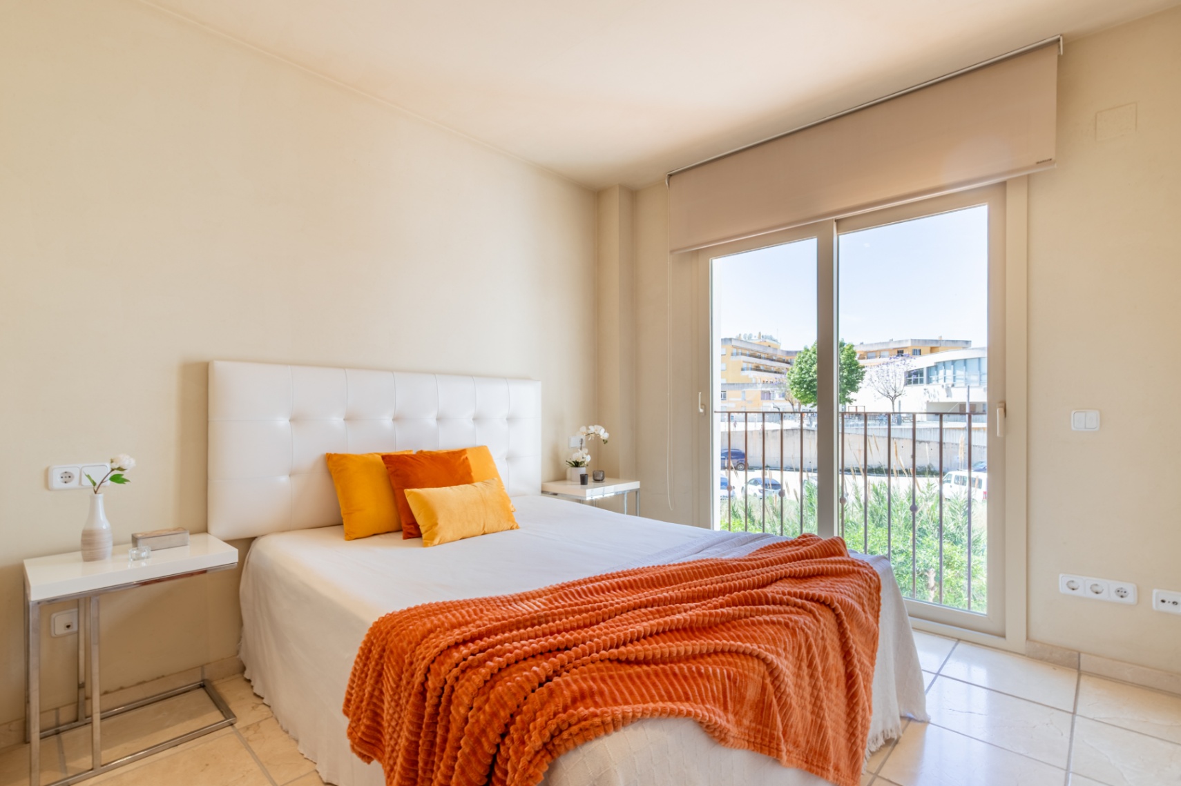 Dormitorio principal con cama doble, cabecero tapizado y suelo de baldosas claras. Acceso directo a un balcón con vistas urbanas.
