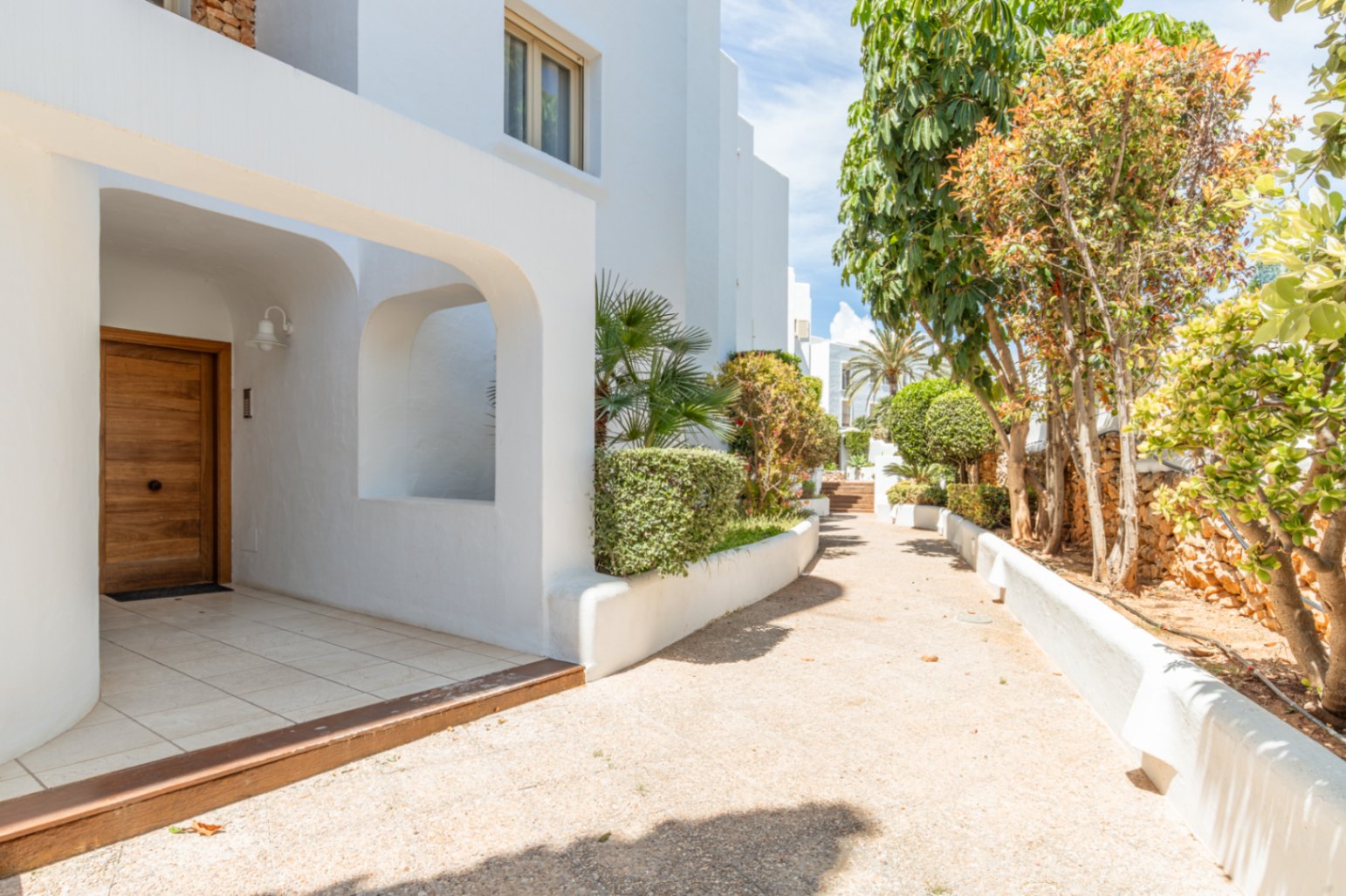 Entrada principal con puerta de madera moderna y fachada blanca arqueada. Un sendero ajardinado con vegetación mediterránea conduce a través del complejo.