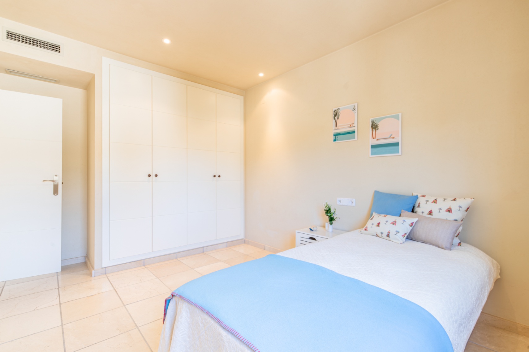 Luminoso dormitorio con amplios armarios empotrados blancos, suelo de baldosas claras y una cama confortable con decoración temática costera. Iluminación empotrada.