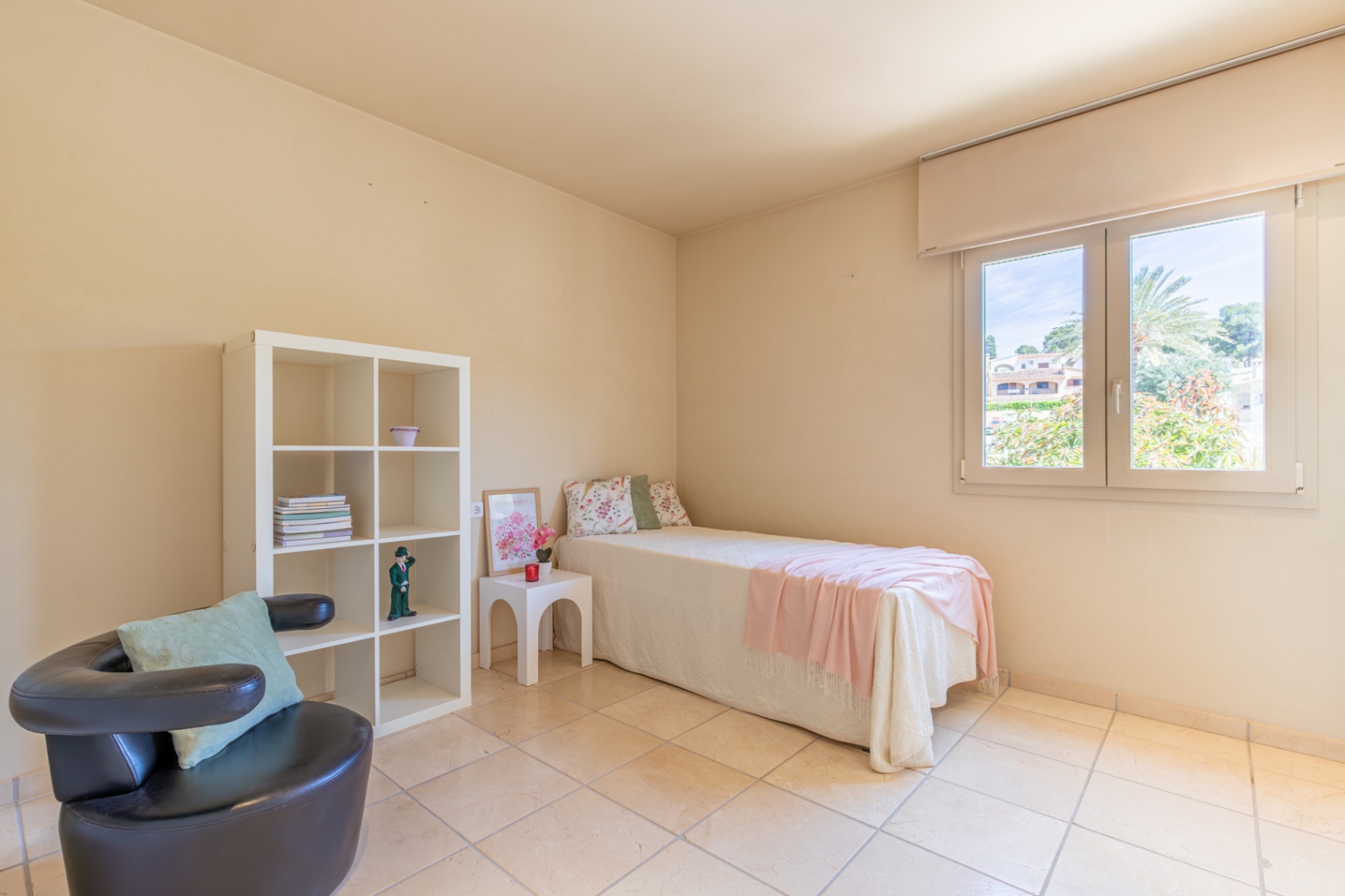 Luminoso dormitorio secundario con cama individual, estantería blanca y sillón oscuro. La ventana ofrece vistas a la vegetación exterior y abundante luz natural sobre el suelo de baldosas claras.