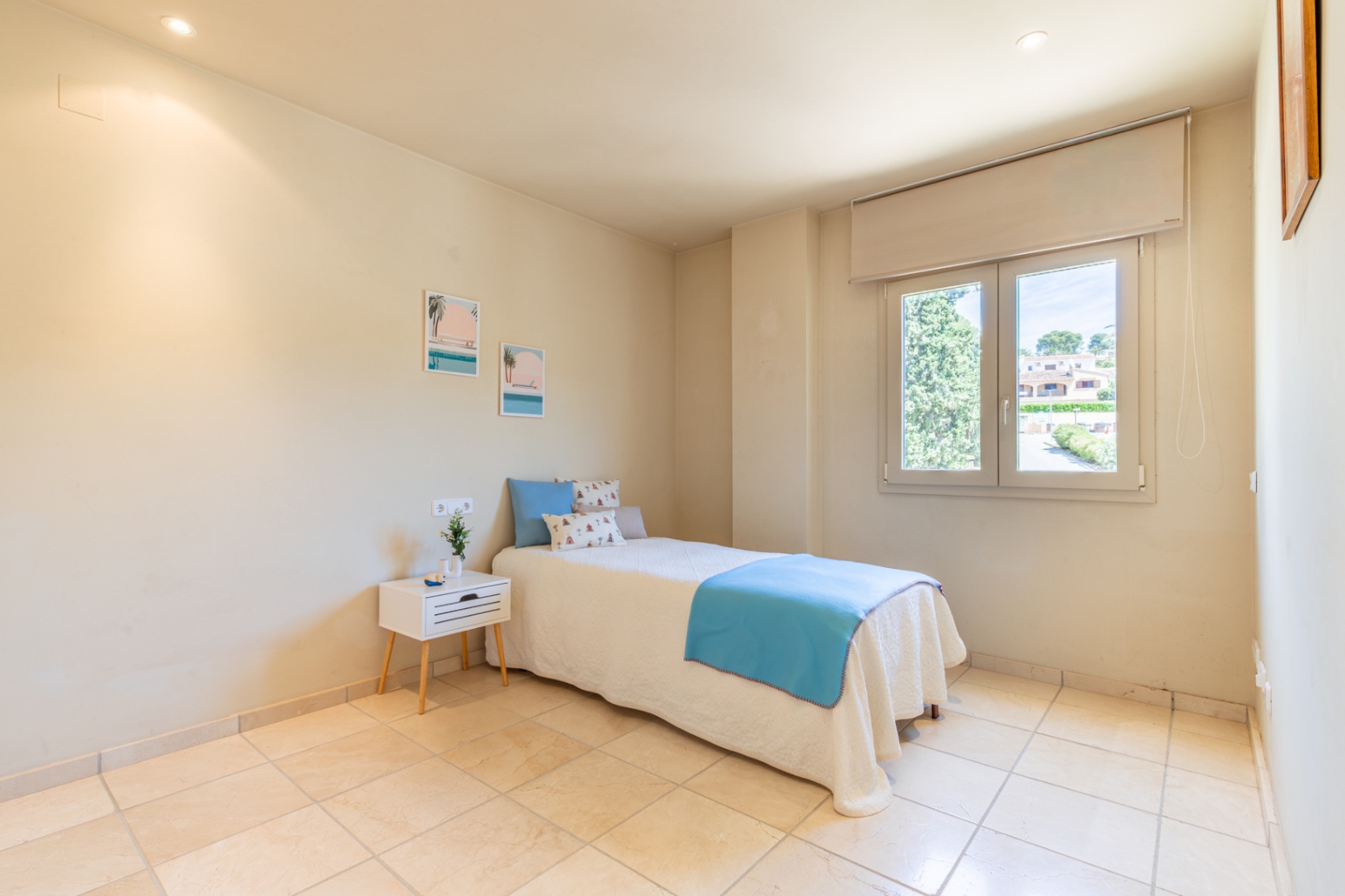 Luminoso dormitorio secundario con cama individual vestida en tonos azules y blancos, suelo de baldosas claras y ventana con vistas a la vegetación y viviendas. Iluminación empotrada.