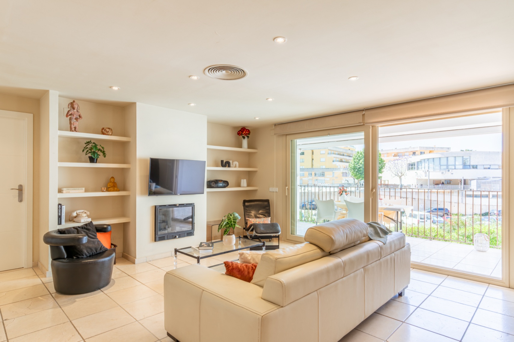 Luminoso salón con sofá crema, suelo de baldosas y estanterías empotradas junto a chimenea y TV. Grandes puertas correderas dan a una terraza privada con vistas urbanas.